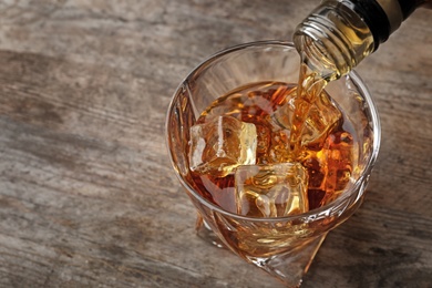 Pouring whiskey from bottle in to glass with ice cubes on table, closeup