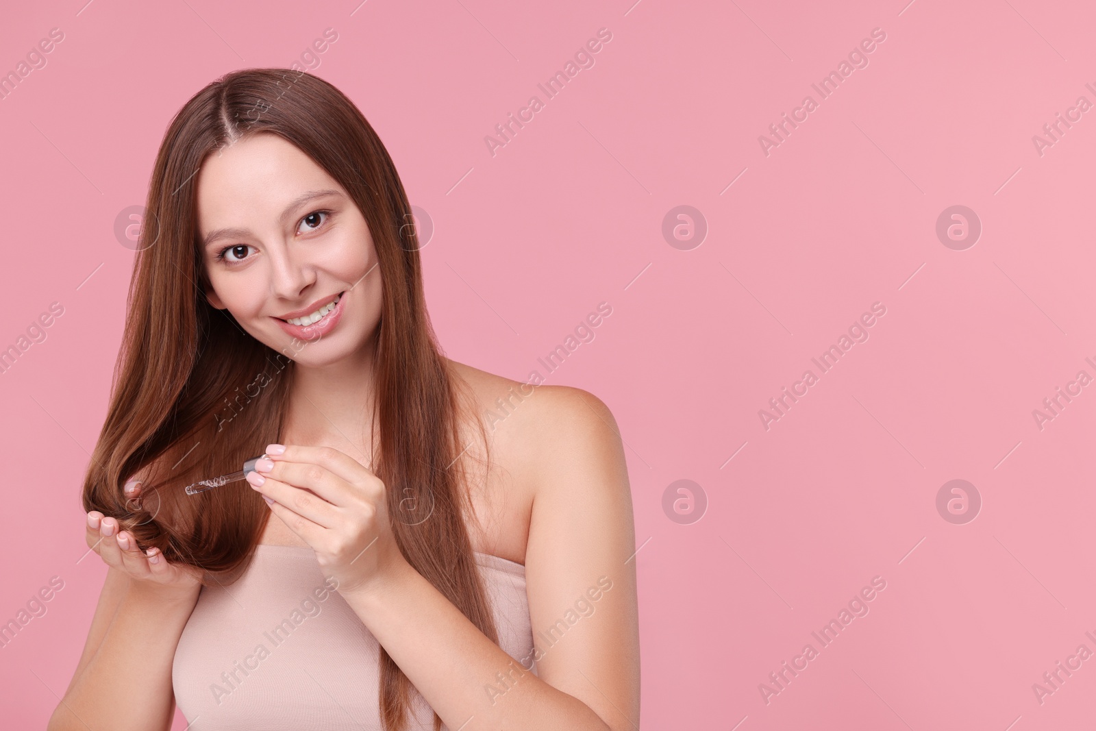 Photo of Beautiful woman applying serum onto hair on pink background, space for text