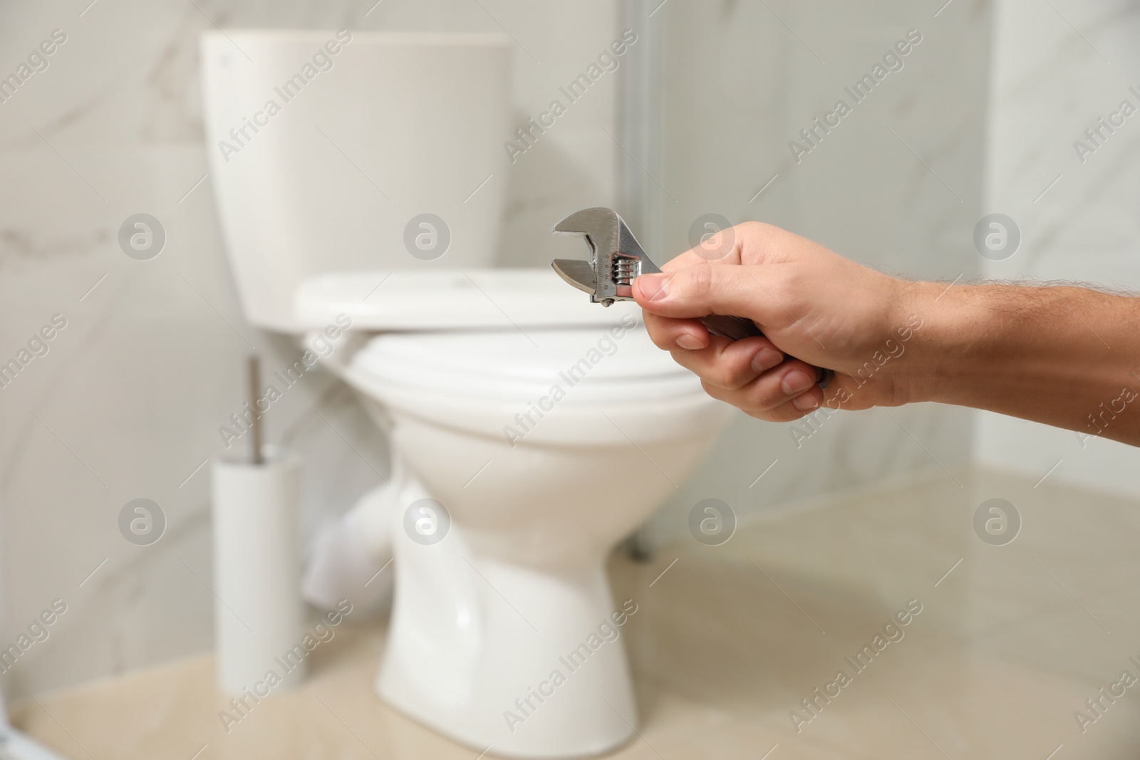 Photo of Professional plumber holding wrench near toilet bowl in bathroom, closeup