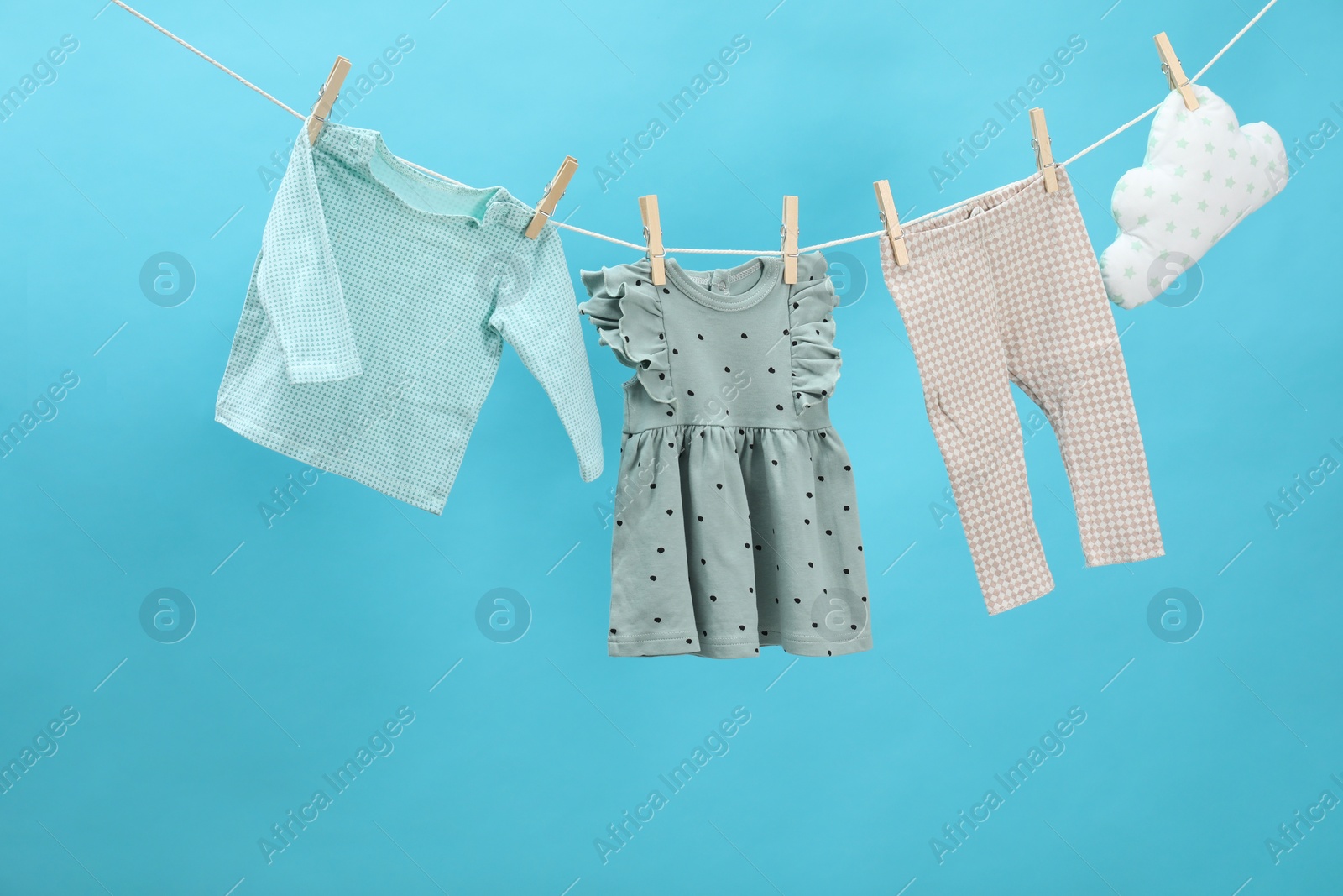 Photo of Different baby clothes and cloud shaped pillow drying on laundry line against light blue background