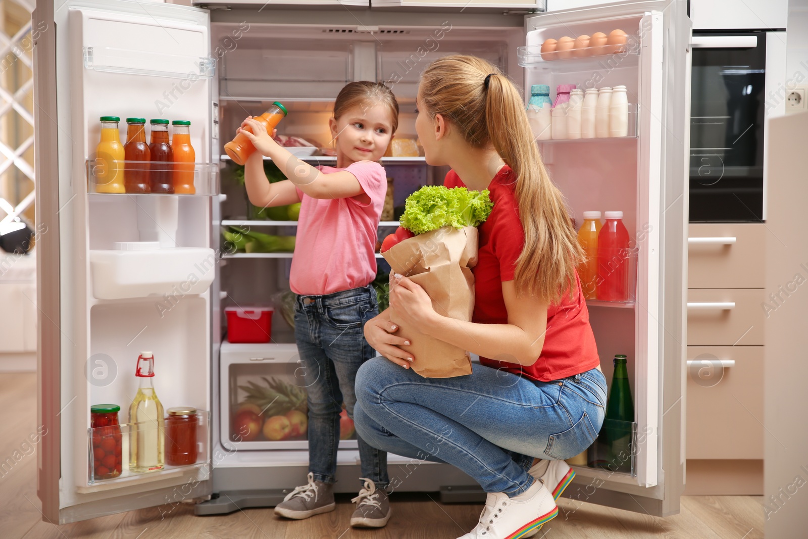 Photo of Young mother with daughter putting food into refrigerator at home after shopping