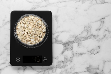 Digital kitchen scale with oat flakes on white marble table, top view. Space for text