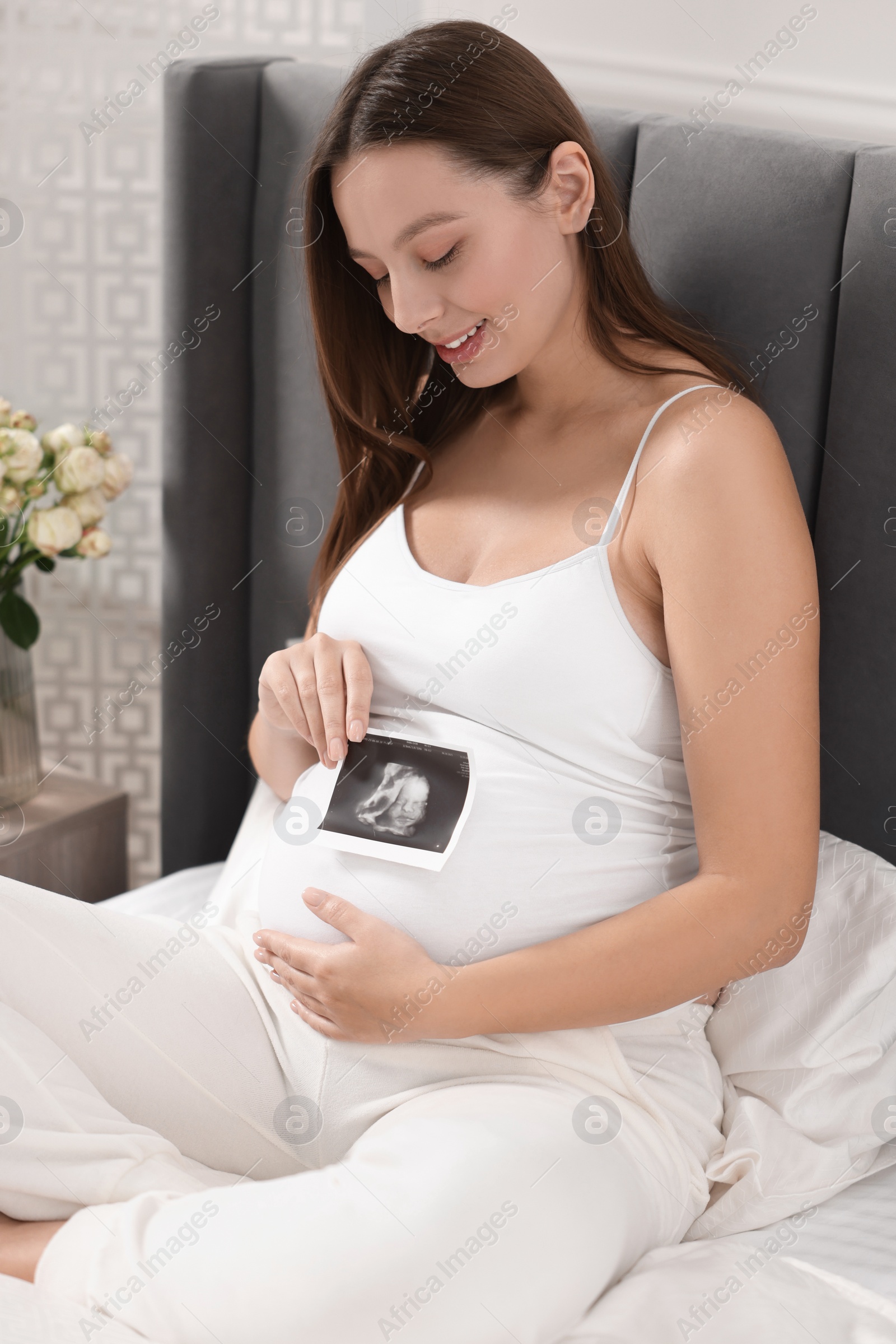 Photo of Pregnant woman with ultrasound picture of baby on bed indoors