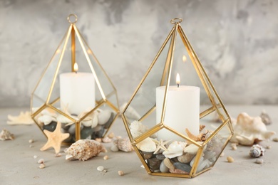 Photo of Stylish glass holders with burning candles, seashells and pebbles on light stone table