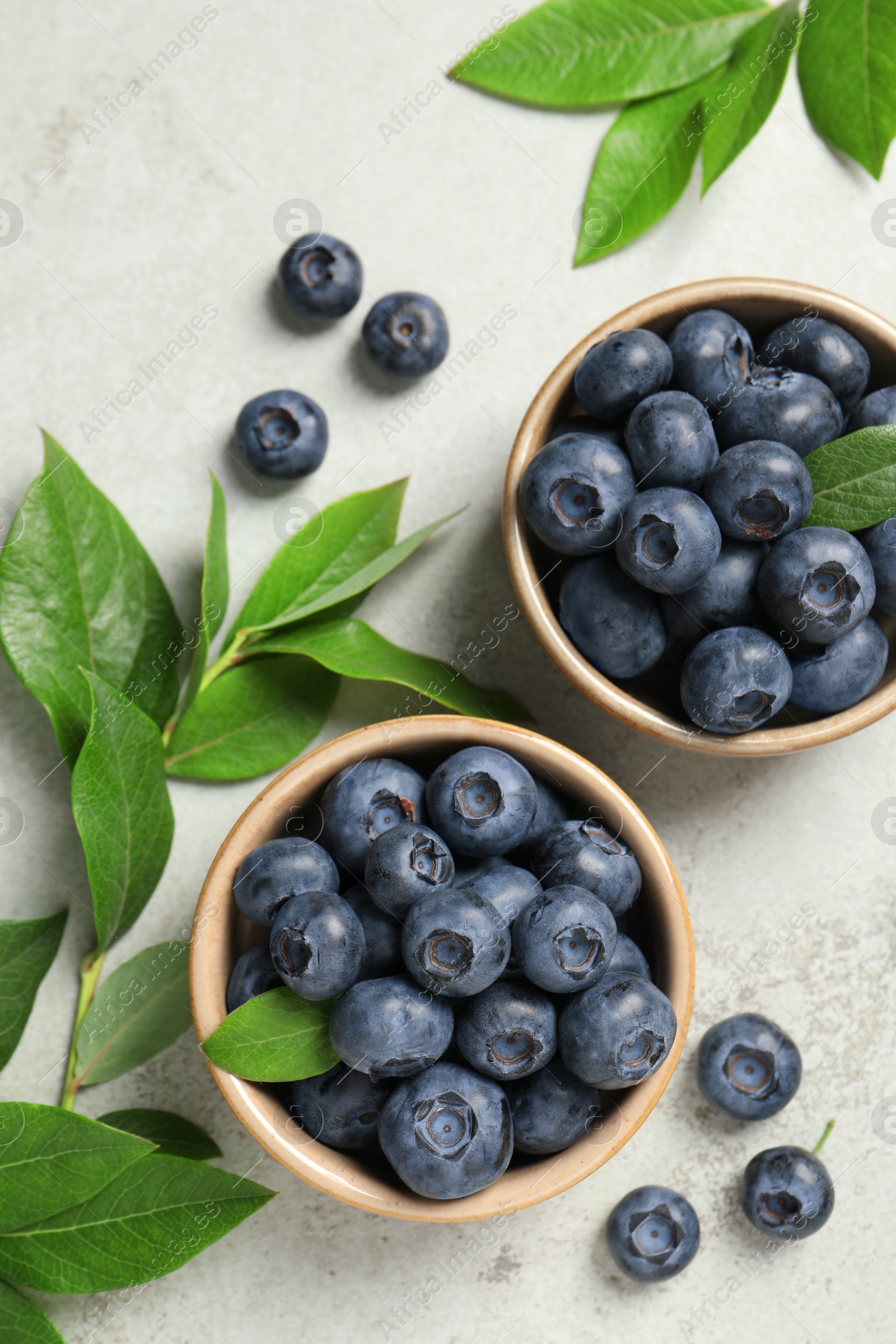 Photo of Tasty fresh blueberries with leaves in bowls on light grey table, flat lay