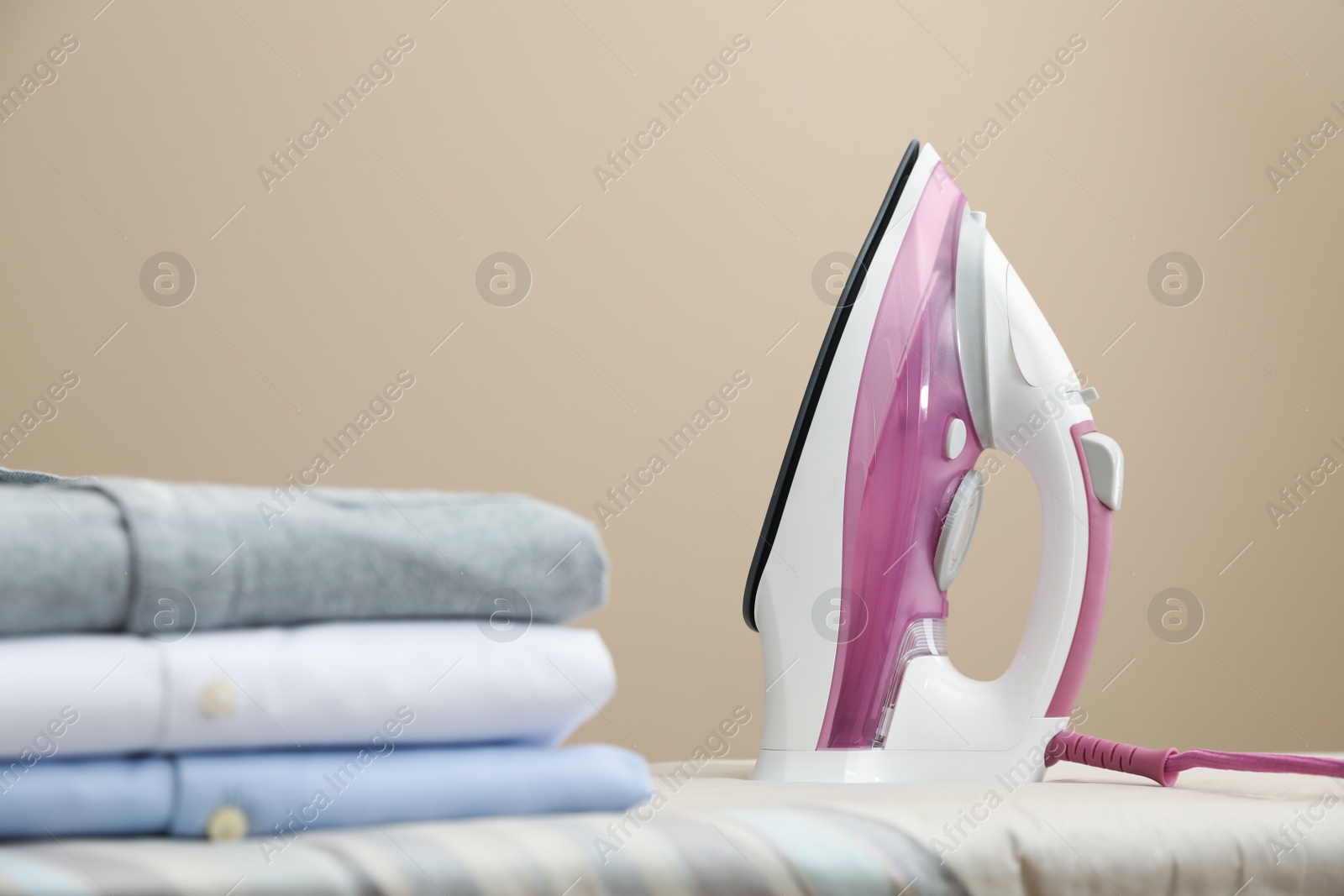 Photo of New modern iron and clean shirts on board against beige background, space for text. Laundry day