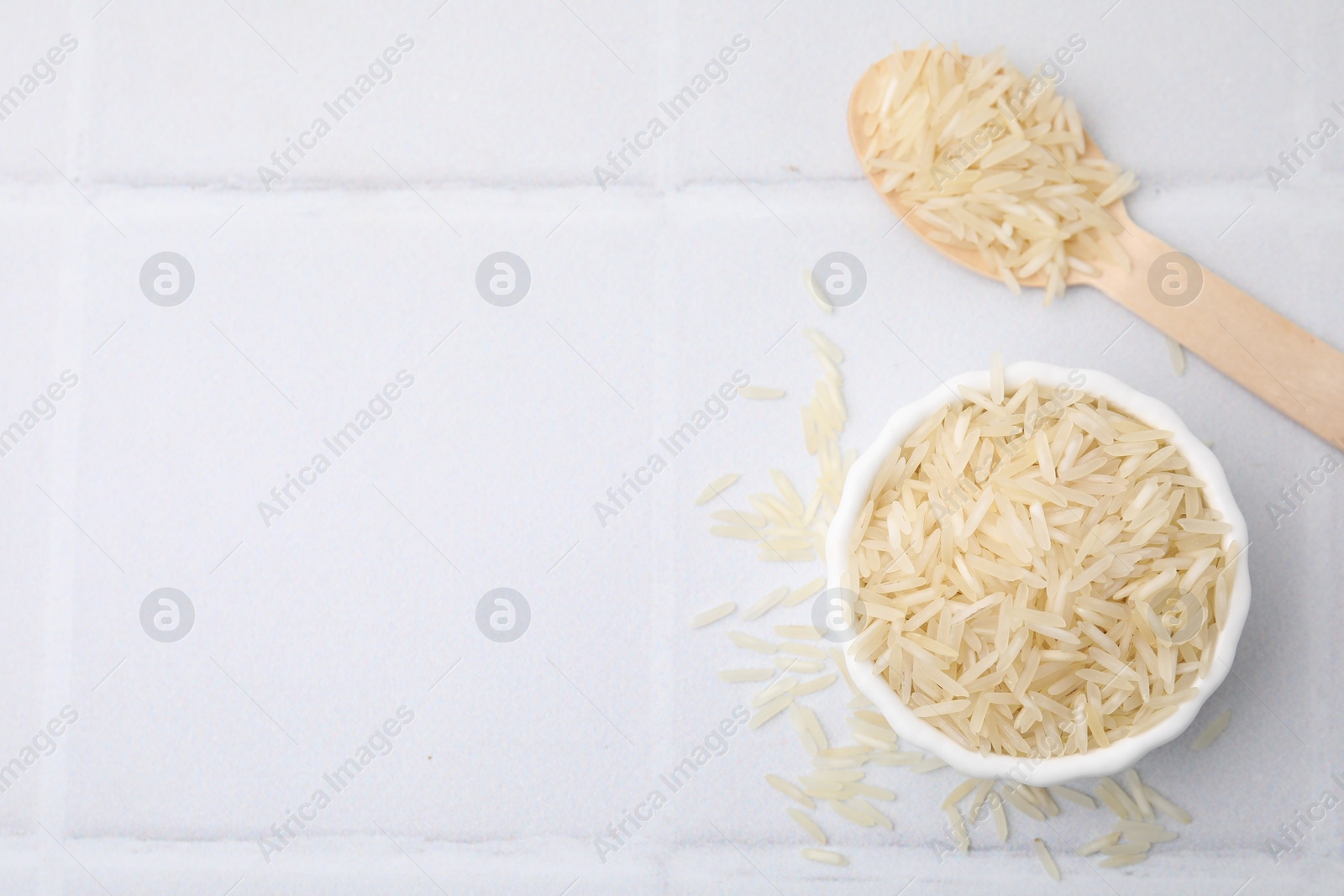 Photo of Bowl and spoon with raw rice on white tiled table, flat lay. Space for text