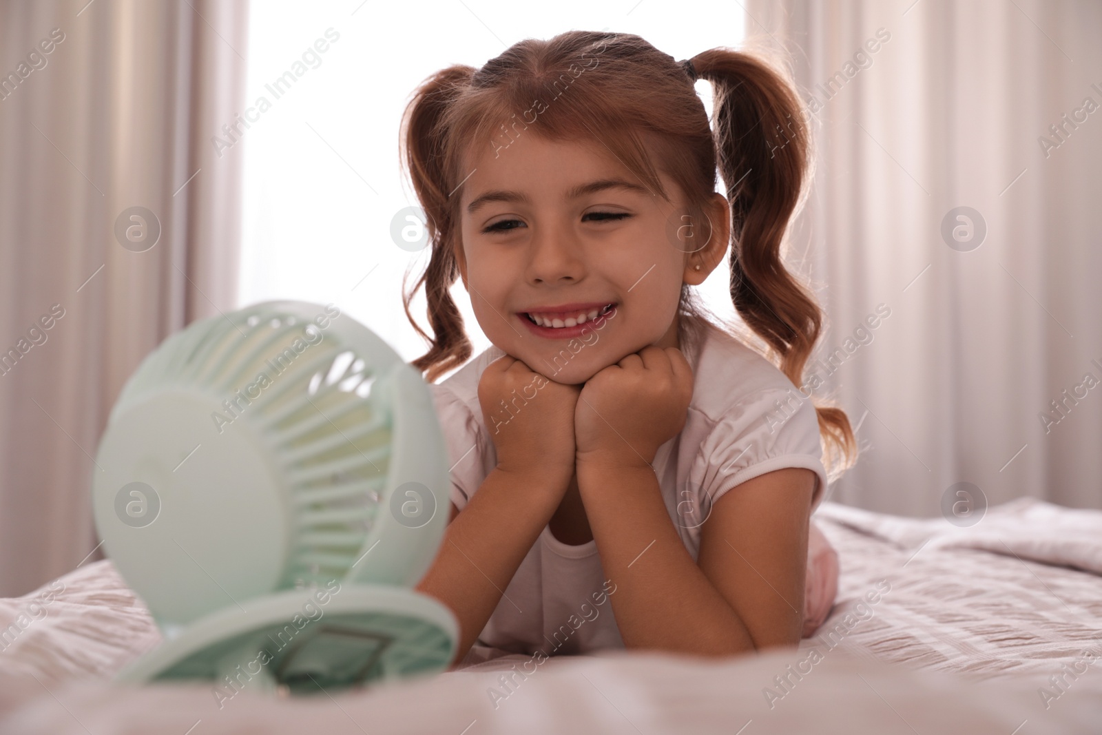 Photo of Little girl enjoying air flow from portable fan on bed in room. Summer heat