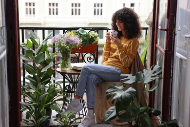 Photo of Young woman with cup of drink relaxing near green houseplants on balcony