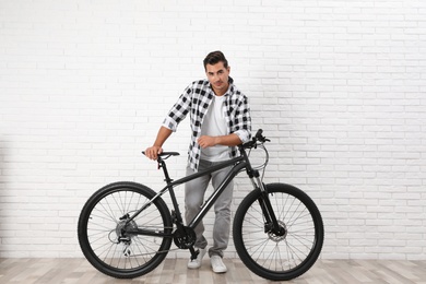 Photo of Handsome young man with modern bicycle near white brick wall indoors