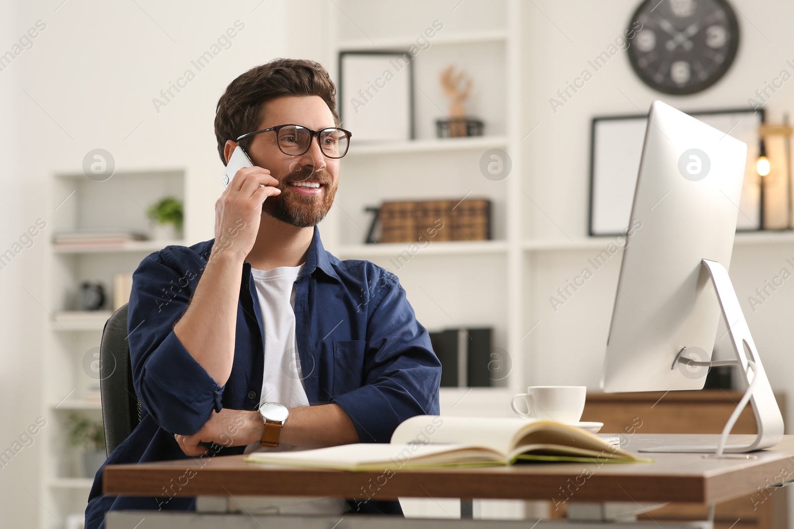 Photo of Home workplace. Happy man talking on smartphone while working with computer at wooden desk in room