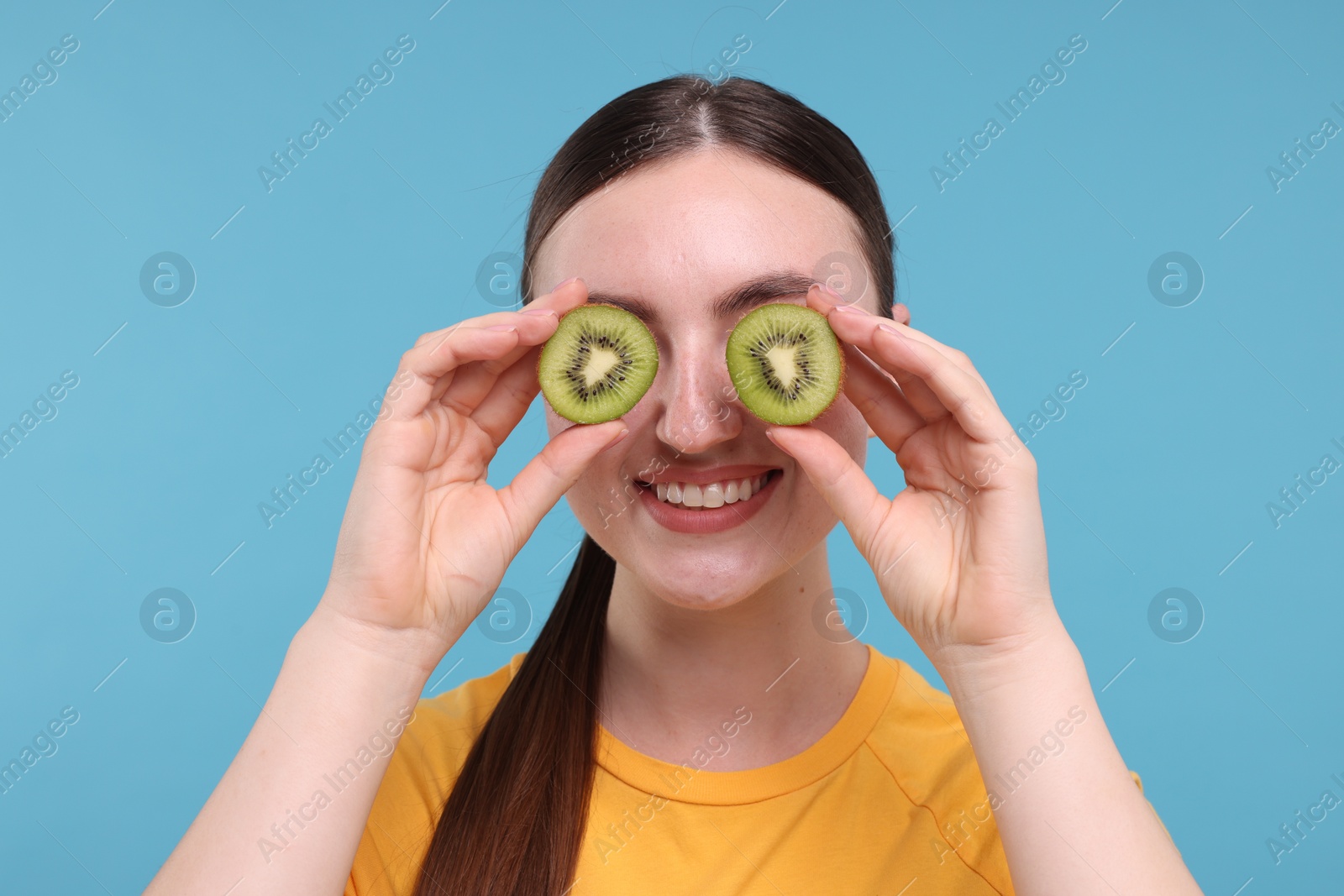 Photo of Smiling woman covering eyes with halves of kiwi on light blue background