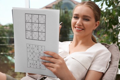 Beautiful young woman solving sudoku puzzle near window indoors, focus on hand