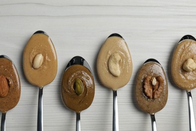 Tasty nut butters in spoons and raw nuts on white wooden table, flat lay. Space for text