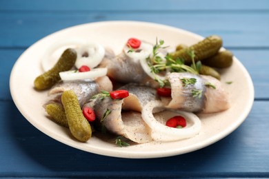 Plate with salted herring fillets, onion rings, thyme, pickles and chili pepper on blue wooden table, closeup