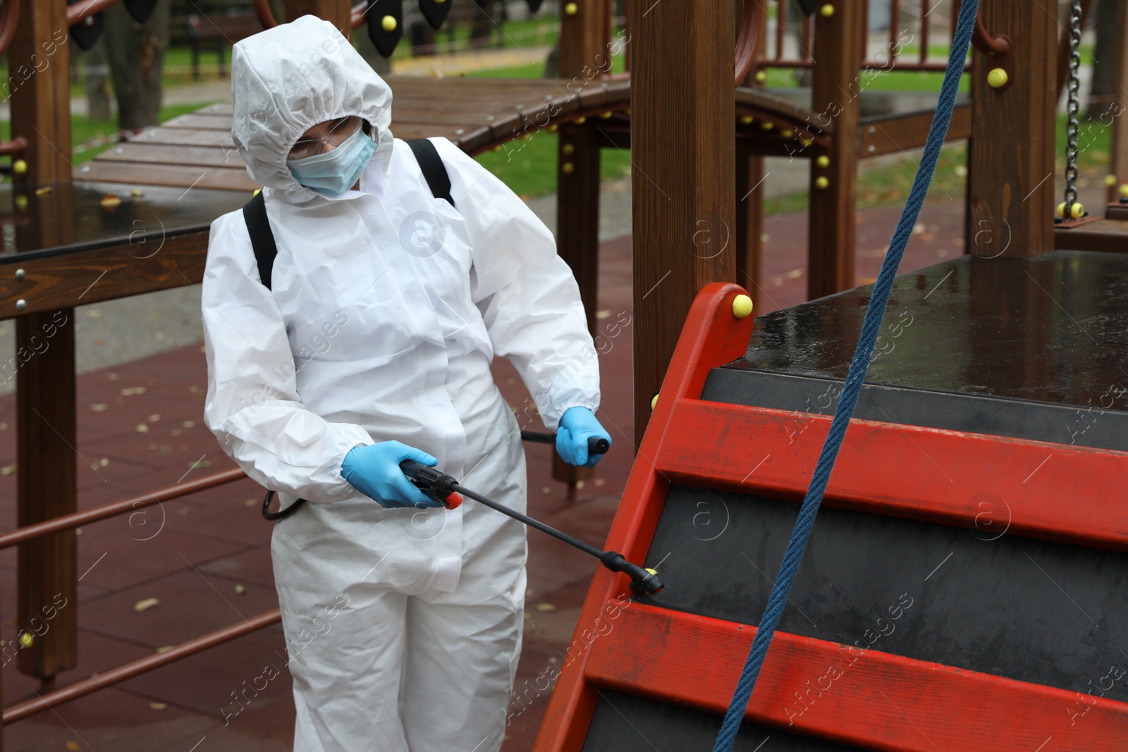 Photo of Woman wearing chemical protective suit with disinfectant sprayer on playground. Preventive measure during coronavirus pandemic