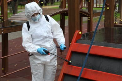 Photo of Woman wearing chemical protective suit with disinfectant sprayer on playground. Preventive measure during coronavirus pandemic