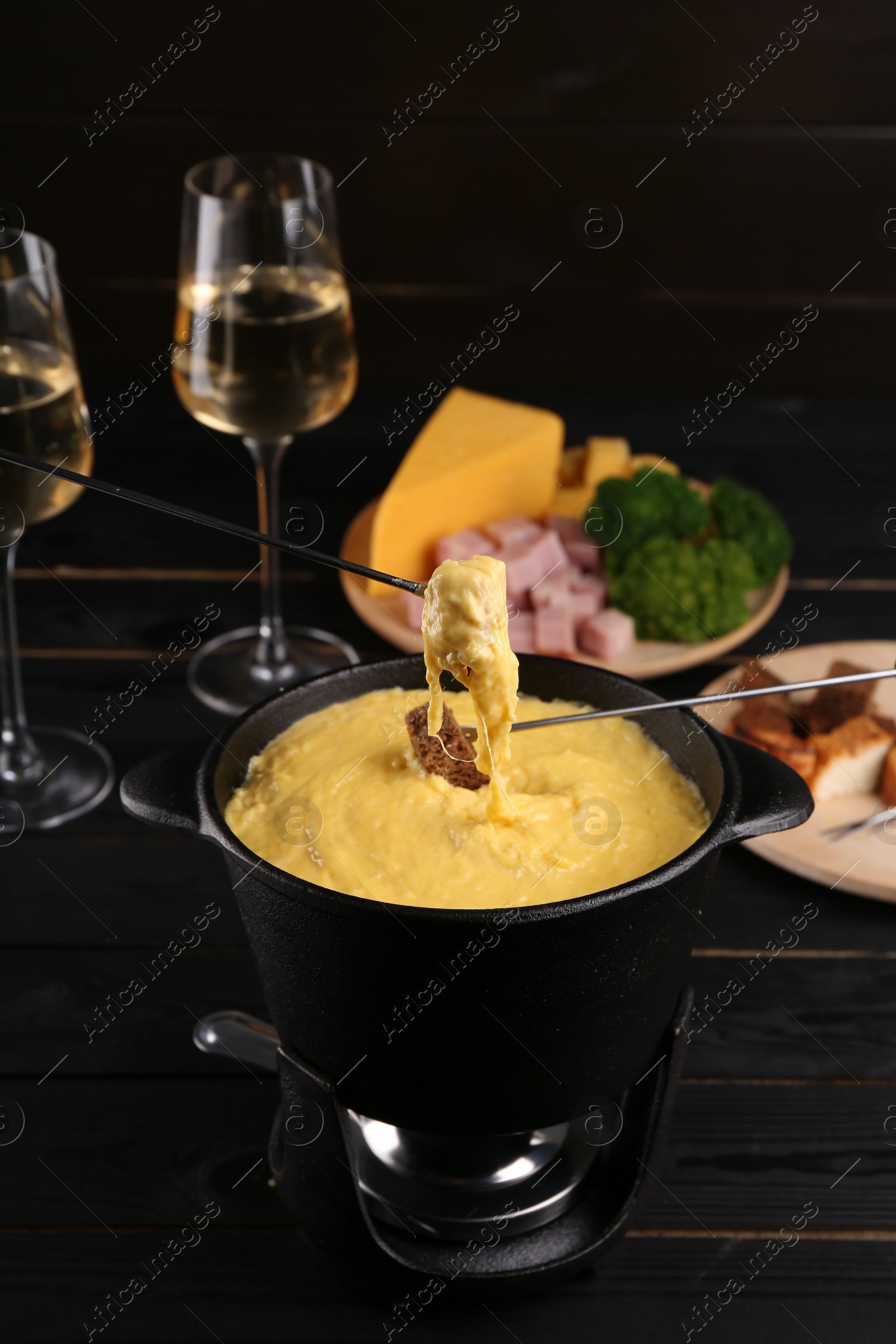 Photo of Dipping pieces of ham and bread into fondue pot with melted cheese on black wooden table