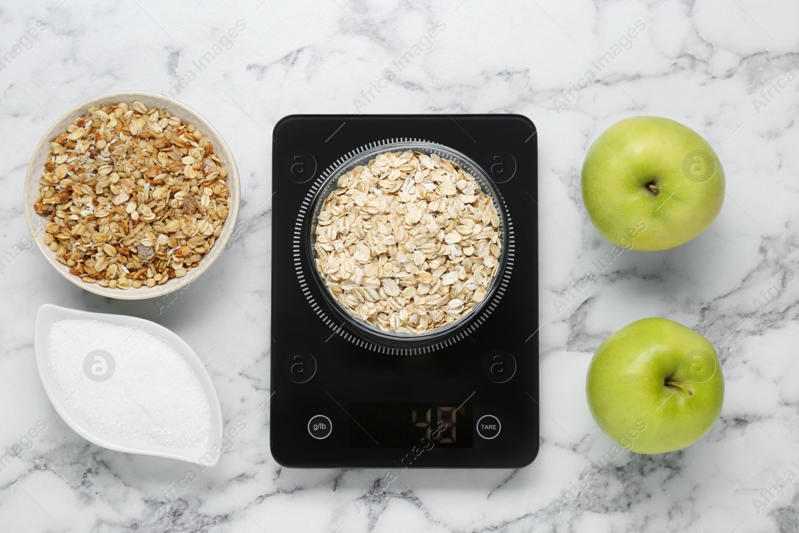 Photo of Digital kitchen scale with oat flakes, granola, apples and sugar on white marble table, flat lay