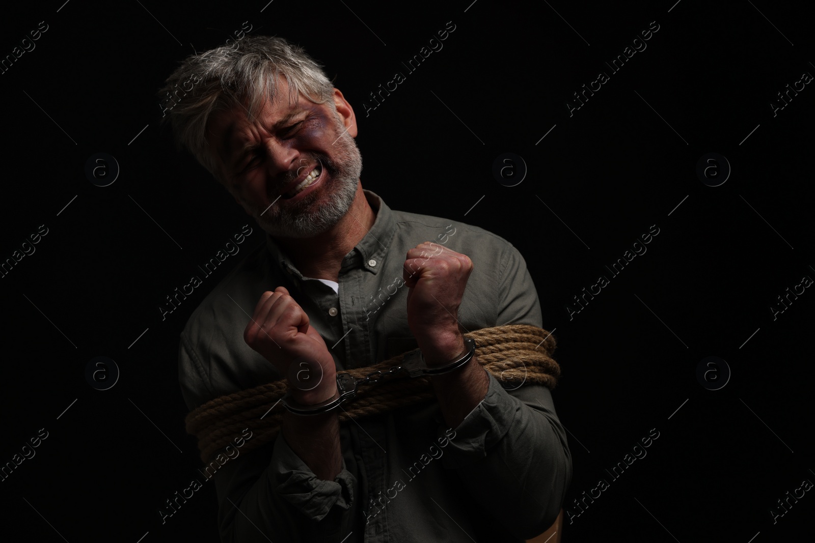 Photo of Emotional beaten man tied with rope on black background. Hostage taking