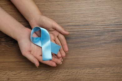 Woman holding light blue awareness ribbon at wooden table, top view. Space for text