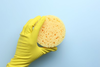 Cleaner in rubber glove holding new yellow sponge on light blue background, top view. Space for text