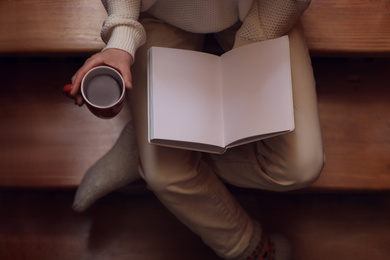 Photo of Woman with cup of coffee reading book on stairs at home, top view