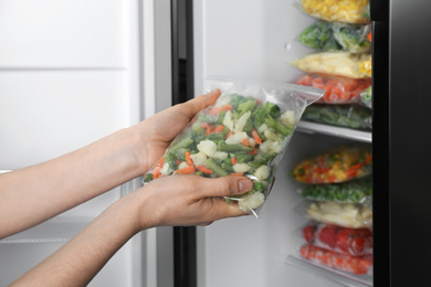 Photo of Woman holding plastic bag with frozen vegetables near open refrigerator, closeup