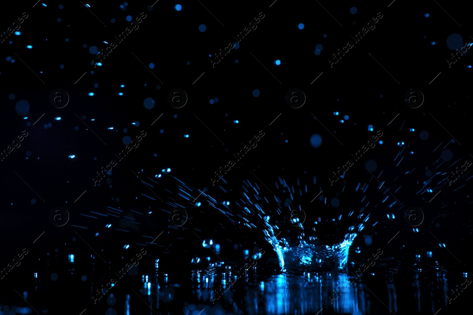 Photo of Rain drop falling down into puddle on dark background, toned in blue