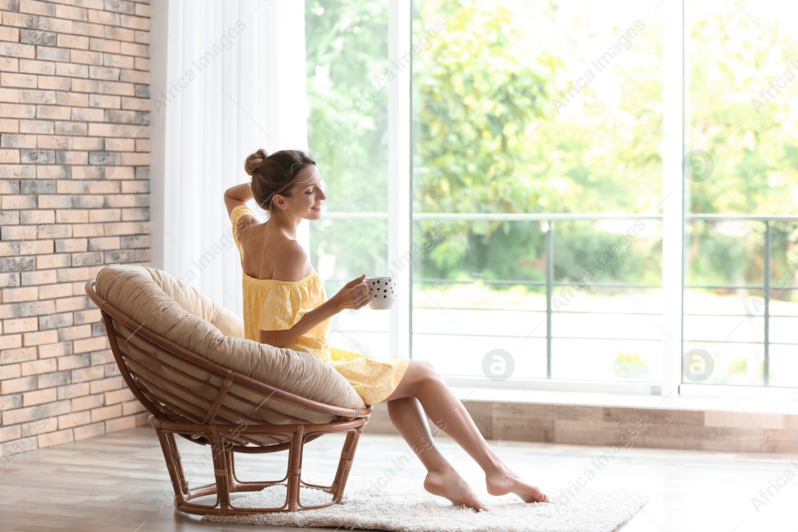 Photo of Young woman with cup of aromatic coffee sitting in papasan chair near window at home