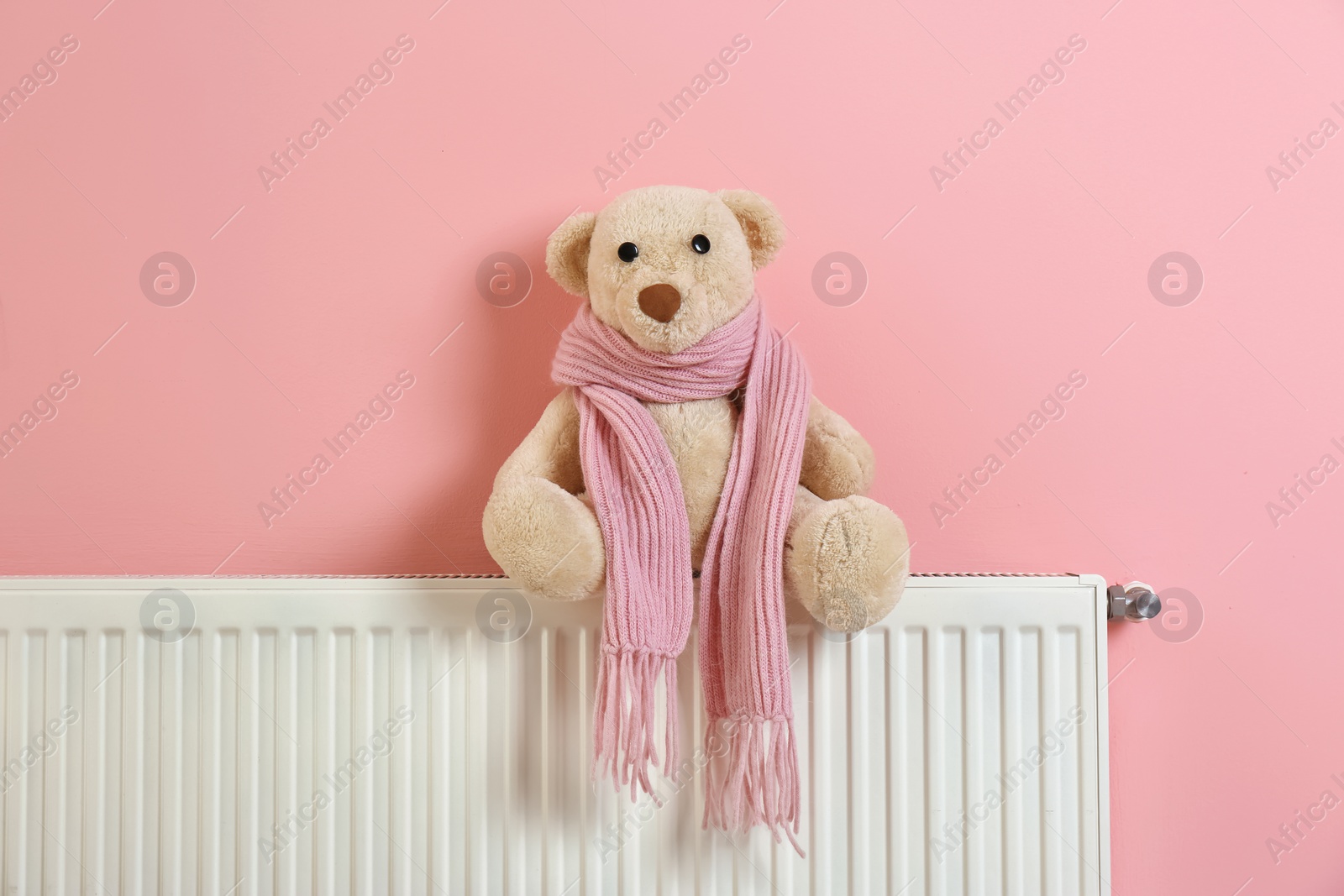 Photo of Teddy bear with knitted scarf on heating radiator near color wall