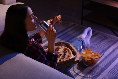 Photo of Young woman drinking from tin can and eating pizza while watching TV in room at night. Bad habit