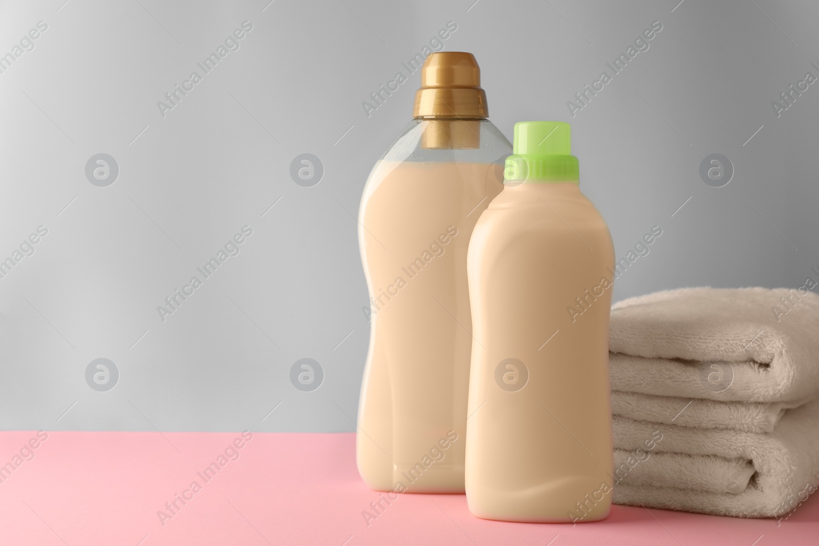 Photo of Bottles of fabric softener and stacked clean towels on pink table against light grey background, space for text