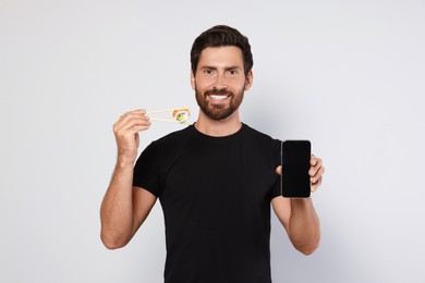 Photo of Happy man holding sushi roll with chopsticks and smartphone on light grey background