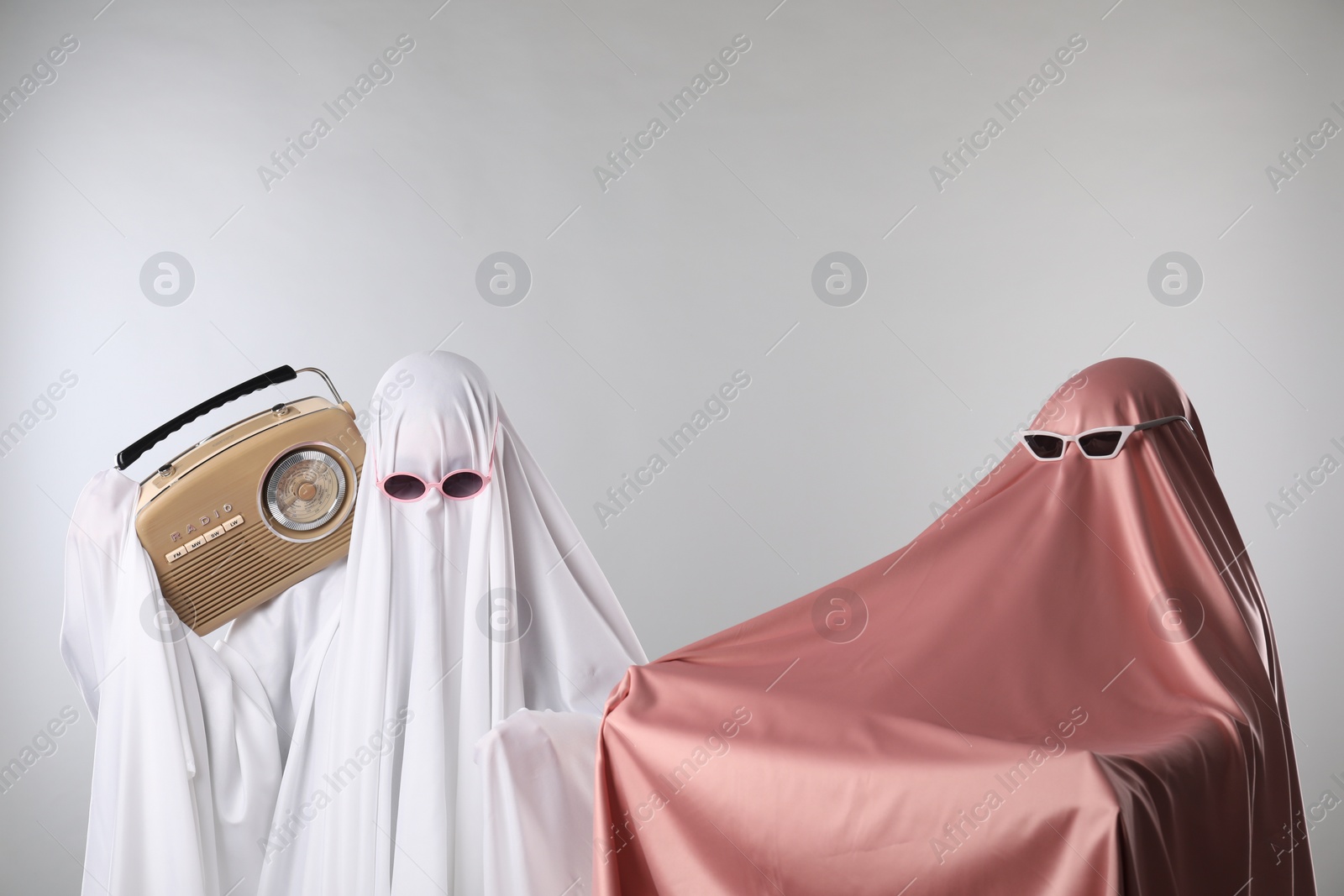 Photo of Glamorous ghosts. Women in color sheets and sunglasses with retro radio receiver on light grey background