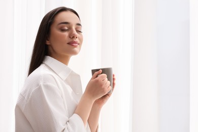 Beautiful woman with cup of drink near window at home, space for text. Lazy morning