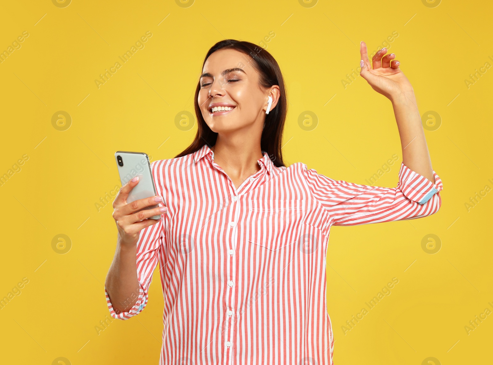 Photo of Happy young woman with smartphone listening to music through wireless earphones on yellow background