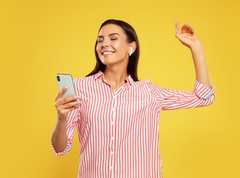 Photo of Happy young woman with smartphone listening to music through wireless earphones on yellow background