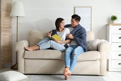 Happy couple covered with warm dark blue plaid reading book on sofa at home