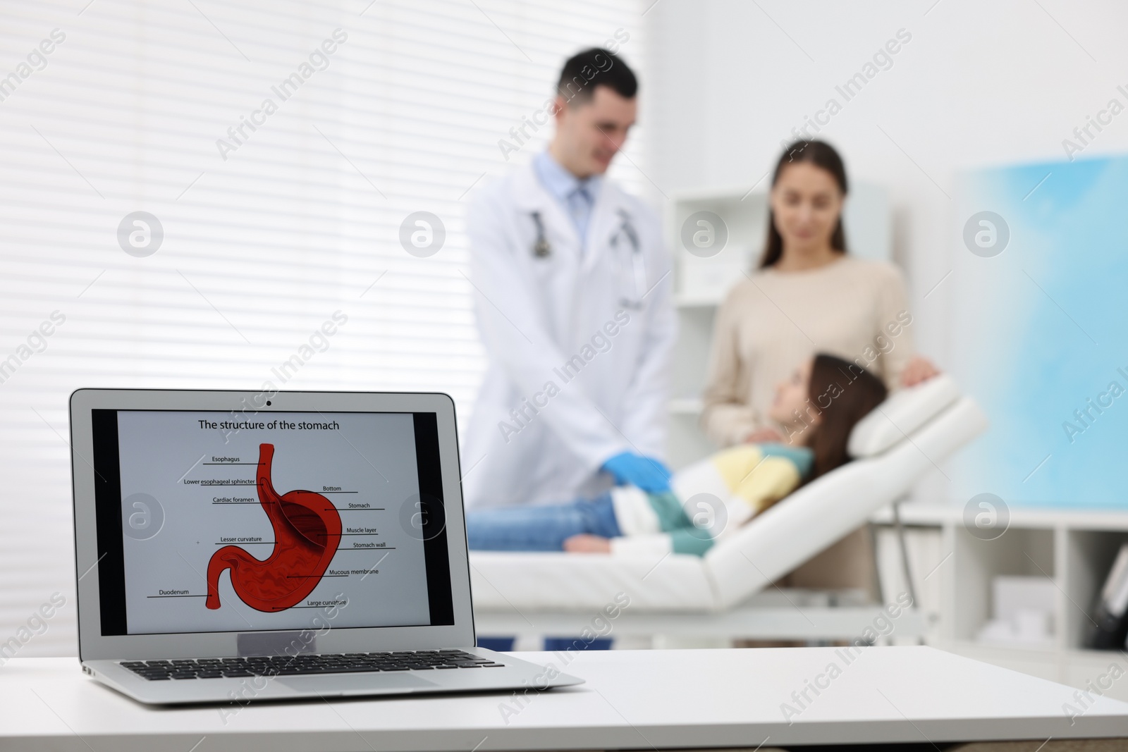 Photo of Gastroenterologist examining girl in clinic, focus on laptop with image of stomach on white table