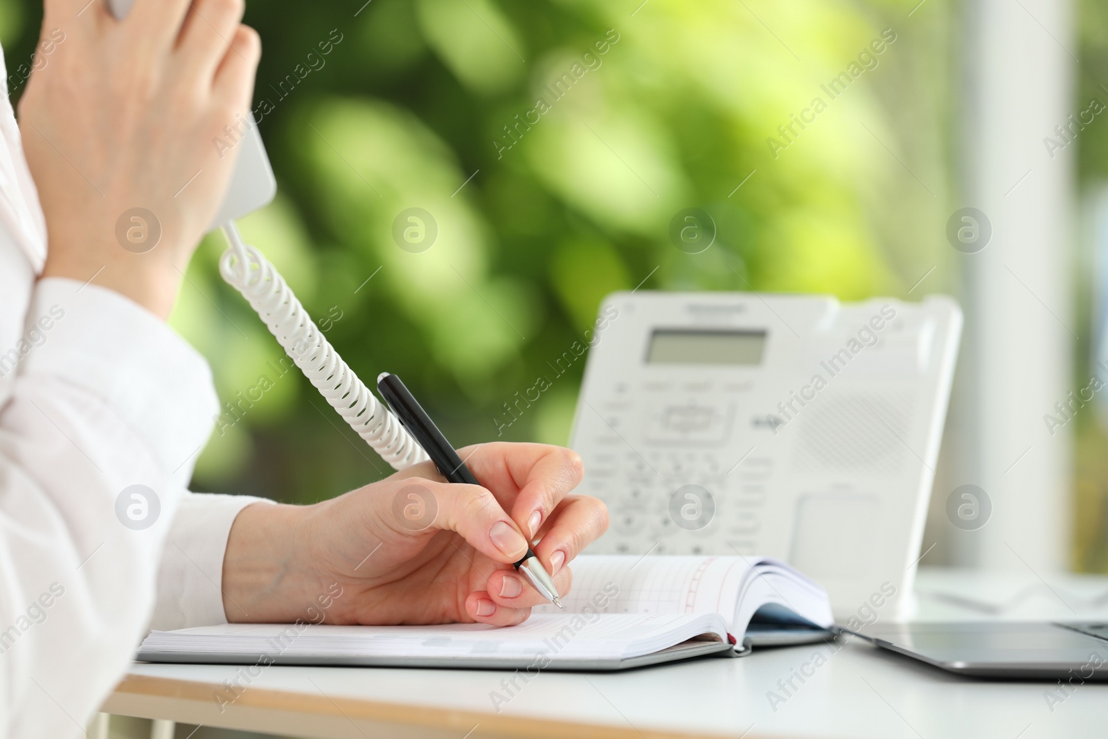 Photo of Assistant with telephone handset writing at white table against blurred green background, closeup