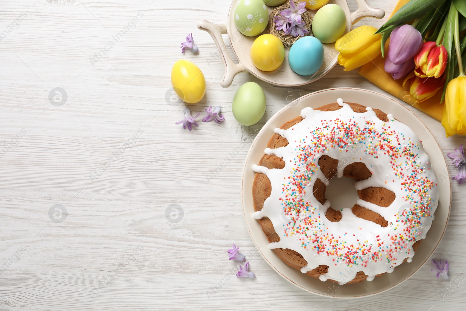 Photo of Glazed Easter cake with sprinkles, painted eggs and flowers on white wooden table, flat lay. Space for text