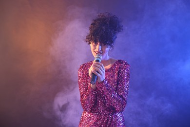 Beautiful young woman with microphone singing on color background in neon lights and smoke