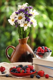 Different fresh ripe berries, beautiful flowers and books on wooden table outdoors