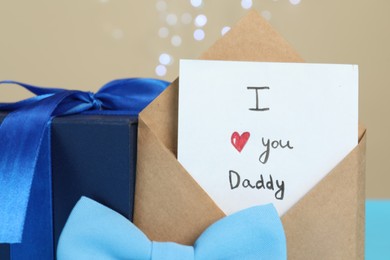 Photo of Happy Father's Day. Card with phrase I Love You, Daddy in envelope and gift box on beige background, closeup