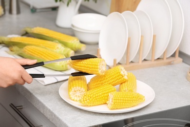 Photo of Woman taking corn cob from plate with tongs in kitchen, closeup