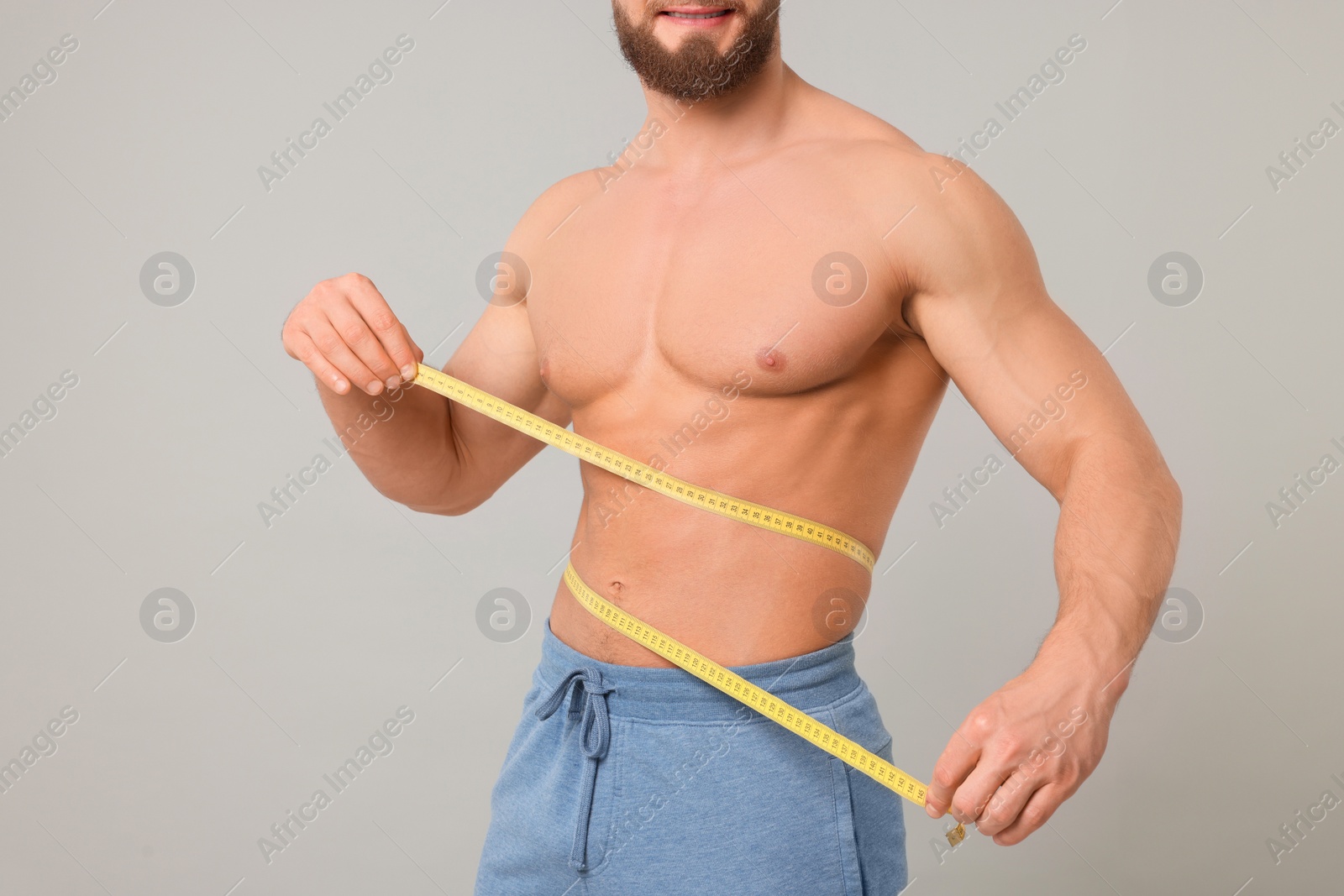 Photo of Athletic man measuring waist with tape on light grey background, closeup. Weight loss concept