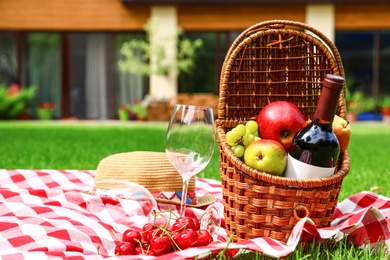 Photo of Picnic basket with fruits and bottle of wine on checkered blanket in garden