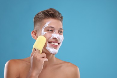 Happy young man washing off face mask with sponge on light blue background. Space for text