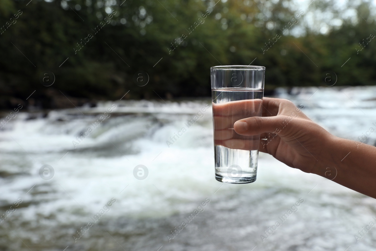 Photo of Woman holding glass of fresh water near river, closeup. Space for text